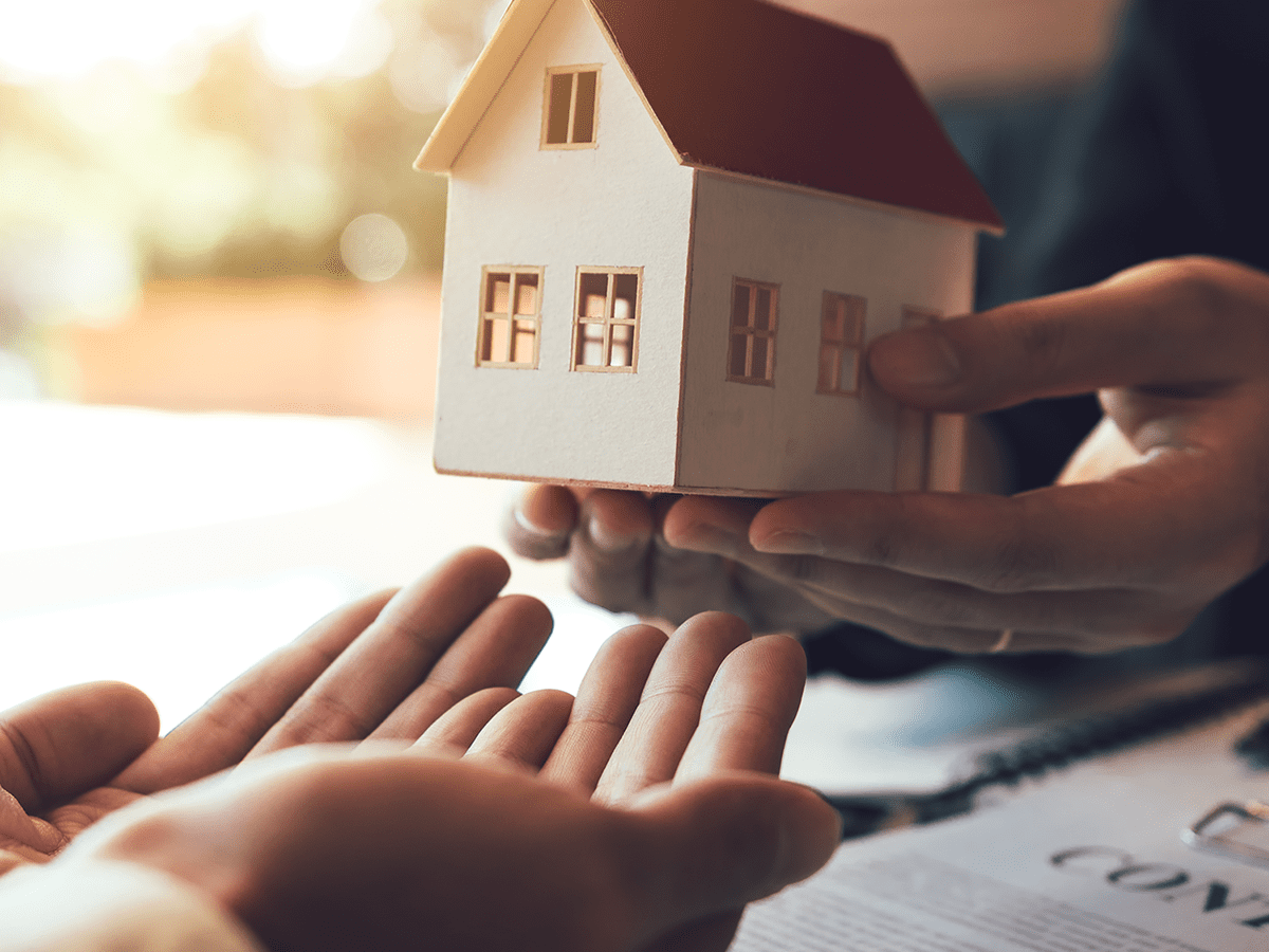 1200x900GMB-4 Person holding a wooden model of a home offering it to another person holding their hands out in front of them